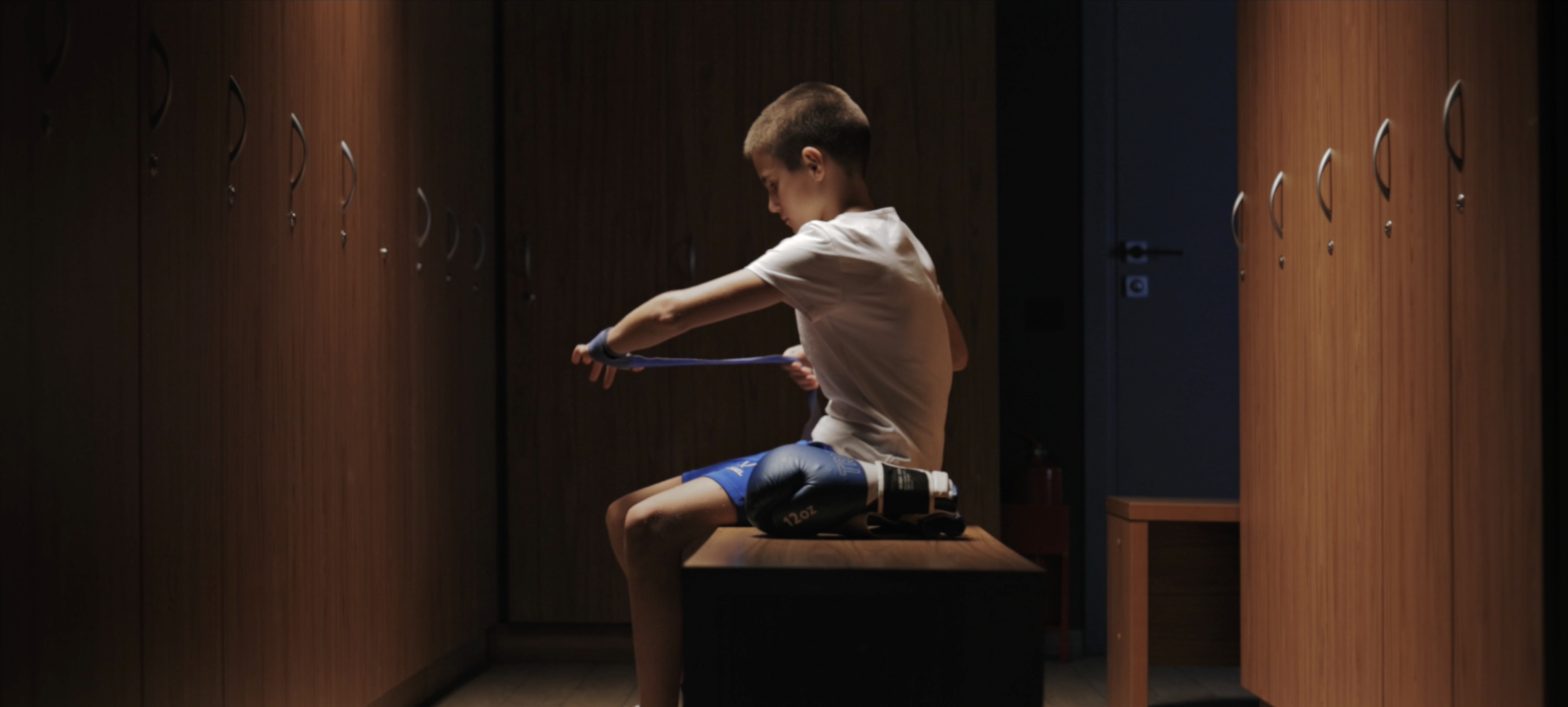 Кадр из имиджевого ролика для ГК ТОЧНО и скульптора Миграна Арутюняна: мальчик готовится к боксерской тренировке в раздевалке — история о преемственности поколений и становлении будущих чемпионов на Аллее славы бокса в Краснодаре